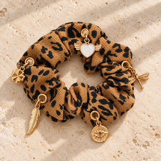 Scrunchie „Wild Leo“ im Leopardenmuster mit Kleeblatt\-, Feder\-, Libellen\-, Augen\- und geflügeltem Herzcharm auf beigem Travertin\.