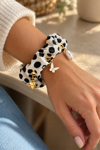 Scrunchie „Black Dots“ mit schwarzen Tupfen und goldfarbenen Herz-, Schmetterlings- und Perlenanhängern, am Handgelenk getragen.