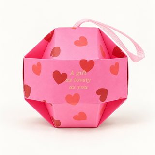 Pinke Geschenkbox in geometrischer Kugelform mit roten Herzen, hellrosa Bandschlaufe und der goldenen Aufschrift „A gift as lovely as you“.