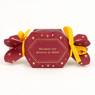 Bordeauxrote Geschenkbox in Bonbonform mit goldenen Sternen, gelben Schleifen und dem Schriftzug „Because you deserve to shine“.