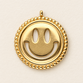 Goldfarbener runder Smiley\-Charm aus Edelstahl mit ausgeschnittenen Augen, lächelndem Mund und perlenartigem Rand auf hellem Hintergrund\.