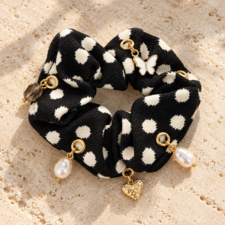 Schwarzer Scrunchie „Cream Dots“ mit cremefarbenen Tupfen sowie Schmetterlings-, Herz- und Perlencharms auf einer beigen Steinfläche.