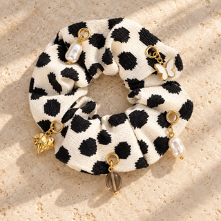 Cremefarbener Scrunchie „Black Dots“ mit schwarzen Tupfen, Perlen-, Herz- und Schmetterlingscharms auf einer beigen Steinfläche.