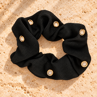 Schwarzer Scrunchie „Classic Black“ mit sechs goldfarbenen Ösen, ohne Charms auf beigem Travertin.