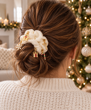 Cremefarbener Scrunchie mit goldenen Ösen und Weihnachtscharms um einen braunen Dutt, im Hintergrund ein beleuchteter Weihnachtsbaum.