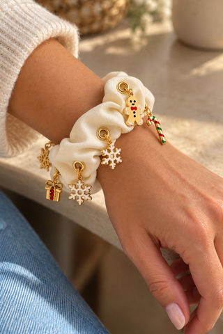 Cremefarbener Scrunchie mit goldenen Ösen und Weihnachtscharms, darunter Lebkuchenmann, Schneeflocken, Geschenk und Zuckerstange, am Handgelenk getragen.