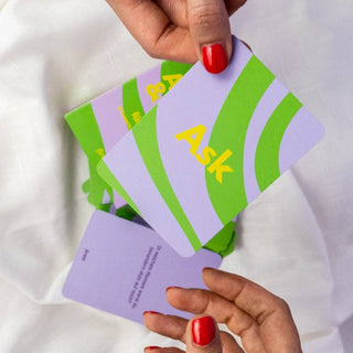 Eine Frau mit rot lackierten Nägeln hält die Ask & Tast Karten in schönem lila und grün in der Hand. 