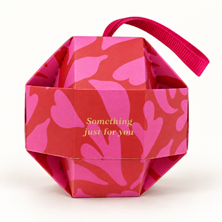 Geometrische Kugel-Geschenkbox mit pink-rotem Muster, magentapinker Bandschlaufe und dem goldenen Schriftzug „Something just for you“.