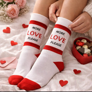 Valentinstagsstimmung weiße Socken mit roten Streien und anderen hell und dunkelroten Details und der Aufschrift More Love please. Die Person sitzt auf dem Bett und in der Umgebung sieht man Pralinen und einen Strauß mit pinken Rosen 