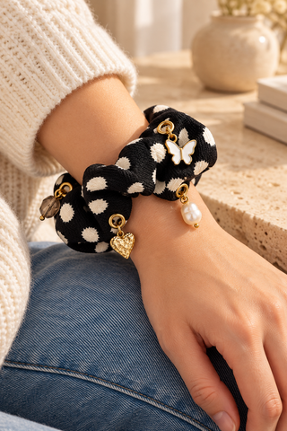 Schwarzer Scrunchie mit cremefarbenen Tupfen, Schmetterlings-, Herz-, Perlen- und braunem Perlencharm, am Handgelenk getragen.