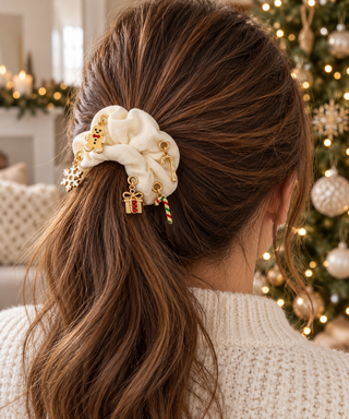Cremefarbener Scrunchie mit goldenen Ösen und Weihnachtscharms in einem braunen Pferdeschwanz vor einem unscharfen Weihnachtsbaum.