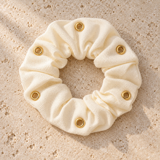 Cremefarbener Scrunchie mit sechs goldenen Ösen ohne Anhänger, flach auf einer hellen Steinoberfläche liegend.
