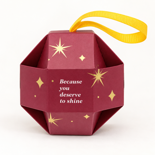 Geometrische bordeauxrote Kugel-Geschenkbox mit goldenen Sternen, gelber Bandschlaufe und der Aufschrift „Because you deserve to shine“.